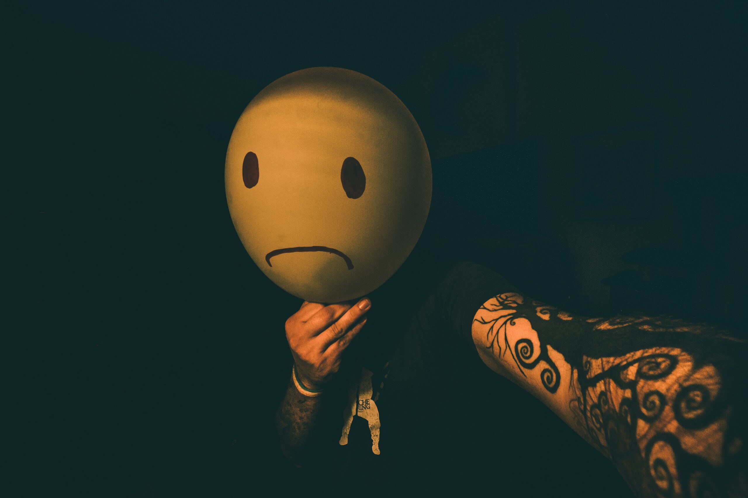 Man holding a balloon with a sad face drawn on it to cover his own – symbolising hidden emotional pain, suicidal thoughts and self-harm in men