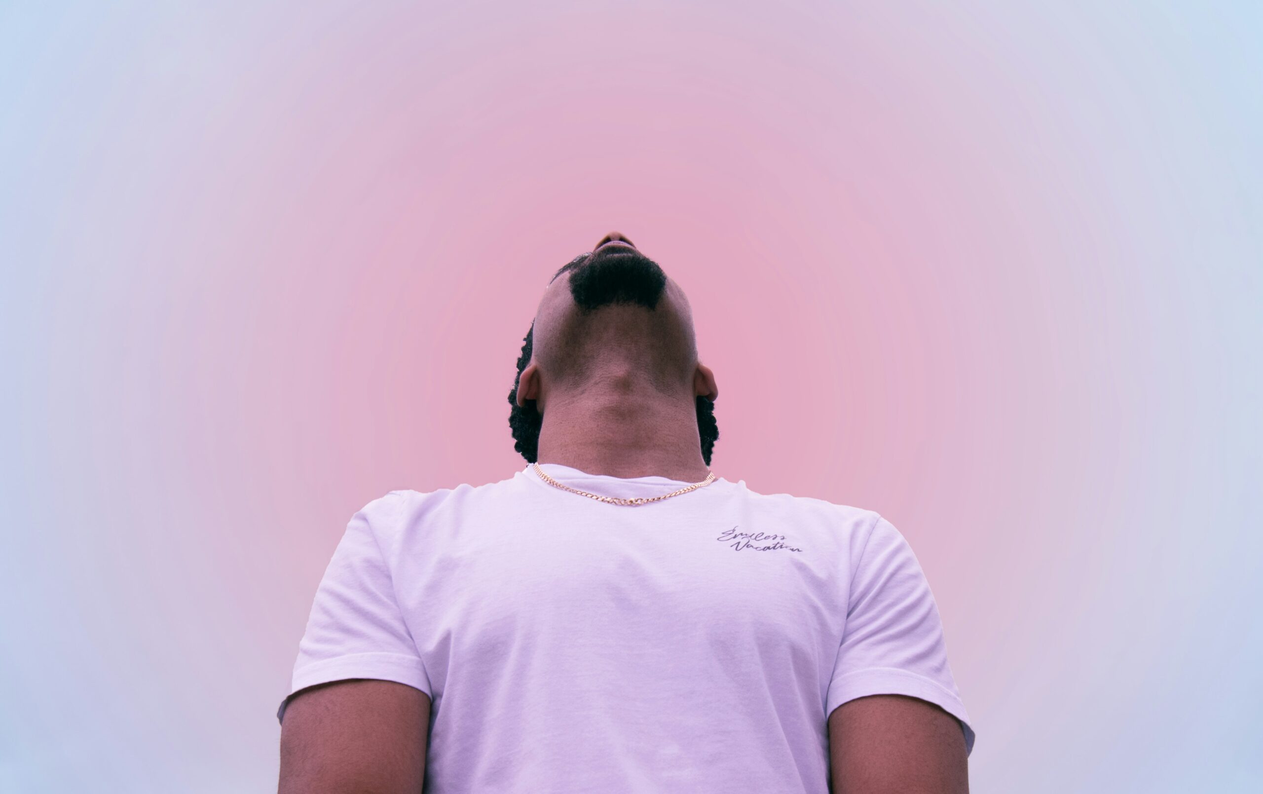 Low-angle shot of a man in a white t-shirt looking upward against a pink and blue gradient sky – representing reflection on identity, self-worth and inner value in men